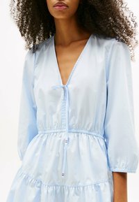 Helles blaues Kleid aus glattem Stoff, mit tiefem V-Ausschnitt, drei Viertelärmel und einem elastischen Taillenbund mit einem rüschengeschichteten Rock.