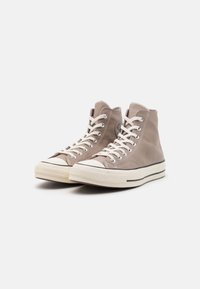 Wysokie trampki ze jasnobrązowej skóry z kremowymi gumowymi podeszwami, białymi sznurówkami i kontrastowym czarnym paskiem wzdłuż podeszwy.