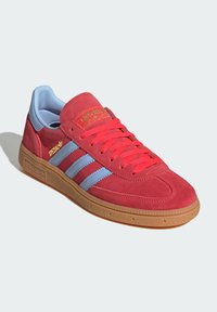 Červené kožené tenisky Adidas Spezial s svetlomodrými pruhmi, červenými šnúrkami, gumenou podrážkou a zlatým brandingom na strane a jazyku.