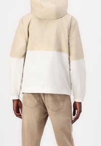 Beżowo-biała kurtka z kapturem, z elastycznymi mankietami i półzamkiem. Lekki, gładki materiał z minimalnym brandingiem na plecach.