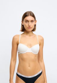 Bílá vyztužená podprsenka s tenkými ramínky, hladkou teksturou, zaoblenými košíčky a jemným švem. Nosená s černými trenýrkami Calvin Klein.