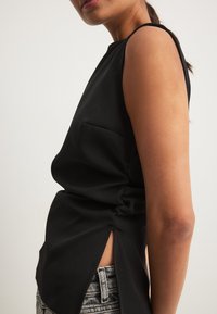 Mustamata ilma varrukat top, millel on küljetasku ja külgmised lõhikud. Sile, kerge kangas, mis on vormitud kujuga, koos naiste teksadega.