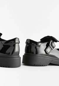 Chaussures Mary Jane en cuir verni noir avec semelles épaisses en caoutchouc et brides à boucle argentée sur fond blanc.