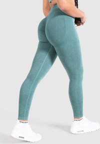 Leggings teal a vita alta con una superficie liscia, caratterizzati da una cucitura a contrasto e abbinati a scarpe da ginnastica bianche.