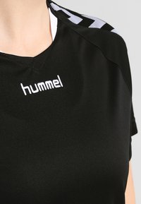 Svart atletisk tröja med korta ärmar, v-ringning och vitt "hummel"-logotyp. Har svarta och vita randiga detaljer på axlarna.
