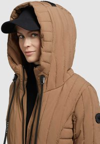 Beige insulated jack met capuchon, voorzien van een gequilt oppervlak, zwarte accenten en een ritssluiting. Gedragen met een zwarte pet eronder.