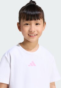 Camiseta blanca de algodón con un pequeño logo rosa de Adidas en el pecho. Cuello redondo y mangas cortas, textura suave.