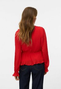 Blusa rossa plissettata con maniche lunghe, vita cintrata e polsini arricciati. Abbinata a pantaloni in denim blu scuro. Design semplice e texture.