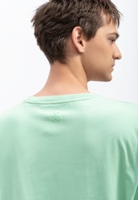 T-shirt vert clair, à manches courtes, texture douce, encolure ronde, avec un logo subtil de ciseaux brodé à l'arrière près du col.
