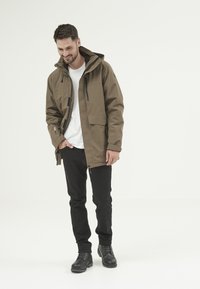 Brun vattentät jacka med huva, två framfickor och dragkedjedetaljer, i kombination med svarta byxor och svarta snörkängor.