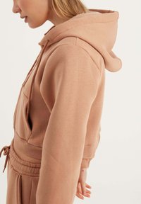 Cappuccio corto rosa con coulisse, tasca frontale e polsini a coste. Il tessuto sembra morbido e ha una texture liscia.