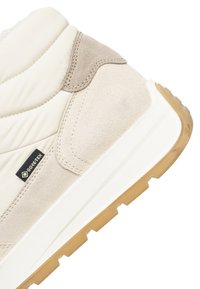 Beige en wit schoenenstuk met suède en doorgestikt textiel, voorzien van een rubberen zool en een zwart Gore-Tex stoffen label.