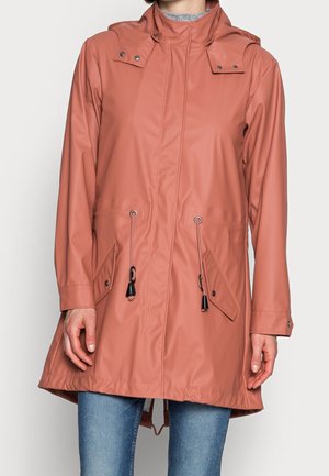 Kobieta w różowym, łososiowym płaszczu przeciwdeszczowym z kapturem, przednimi kieszeniami i ściągaczem w talii, w połączeniu z jasnoniebieskimi dżinsami.