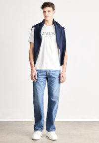 Camiseta blanca con el logo "CALVIN", suéter de punto azul marino sobre los hombros, vaqueros azul rectos y zapatillas blancas. Conjunto sencillo y casual.