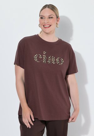 Glimlachende vrouw met licht haar draagt een bruin T-shirt met luipaardprint "ciao" tekst en bijpassende bruine broek tegen een witte achtergrond.