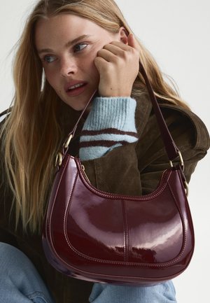 Burgunderfarbene Lederschultertasche mit glänzender Oberfläche, geschwungener Form, doppelten Nähten und einem braunen Trageriemen.