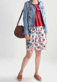 Veste en denim sur un top rouge à volants, associée à une jupe à imprimé floral avec des motifs roses et verts. Sac à bandoulière marron et espadrilles.