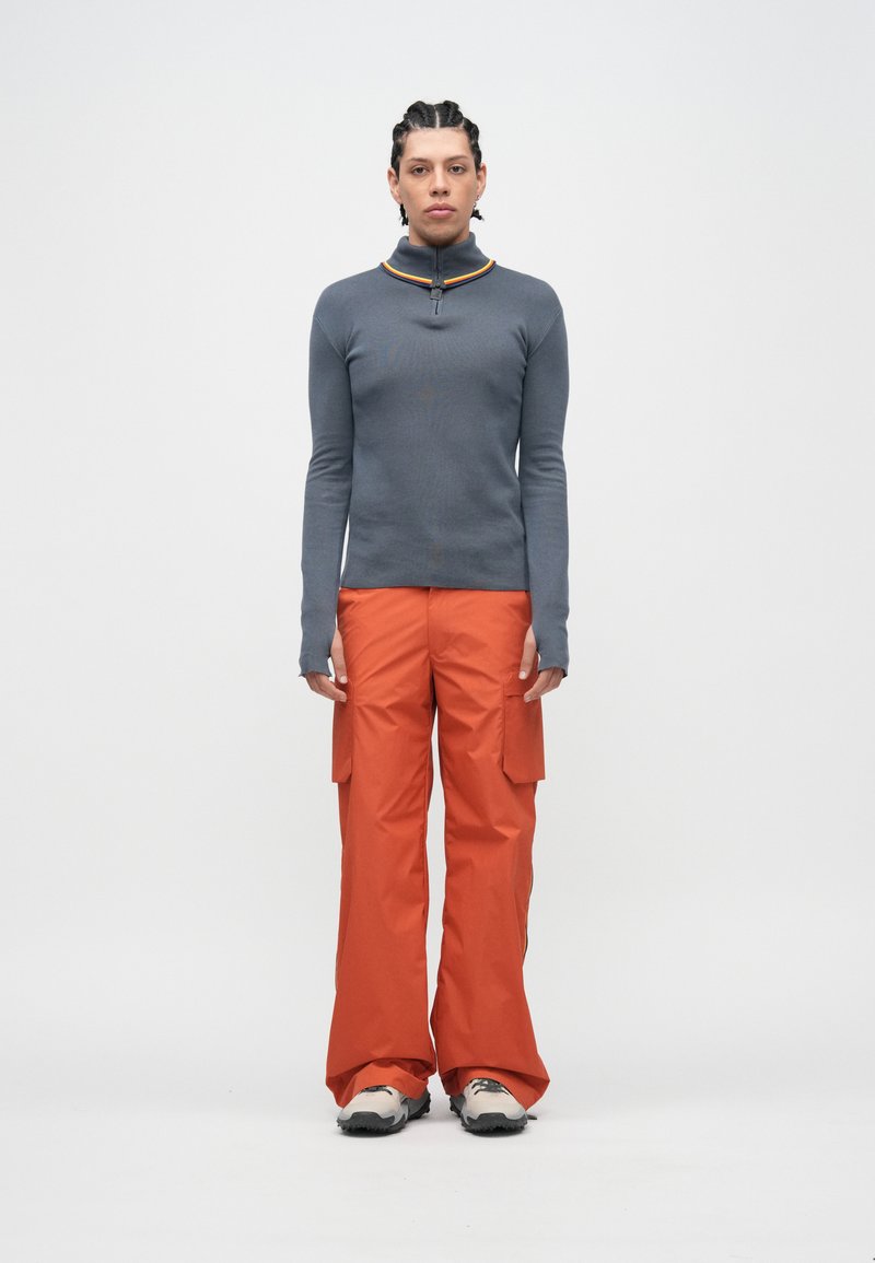 Pelēka garroku jaka ar rāvējslēdzēju un apelsīnu un dzeltenu akcentu, kopā ar spilgti oranžām cargo biksēm ar kabatām un robustiem āra apaviem.