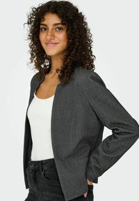 Jeune femme aux cheveux bouclés portant un blazer en chevrons gris sur un haut blanc et un jean noir, souriant avec une main sur la hanche.