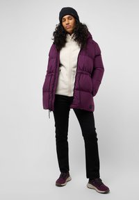 Paarse pufferjack met capuchon, strakke taille en ritssluiting aan de voorkant, gedragen over een crèmekleurige fleece top. Gecombineerd met zwarte broek en paarse sneakers.
