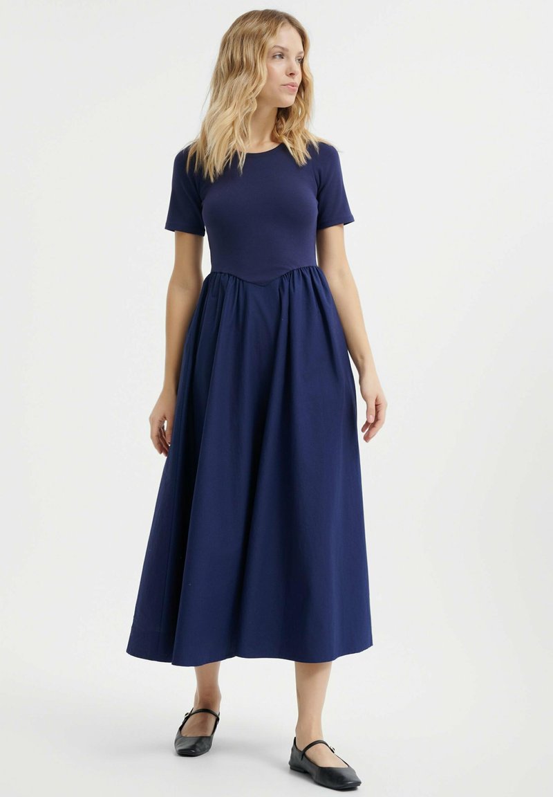 Marineblauwe maxi-jurk met korte mouwen, ronde halslijn, getailleerde bodice en geraffineerde rok. Gepaard met zwarte ballerina's. Zorgvuldig geweven stof.