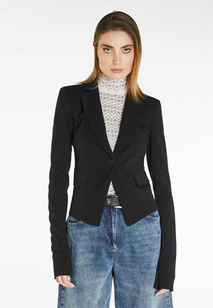 Blazer nero su misura con chiusura a un bottone, colletto a rever e spalle strutturate, indossato sopra un dolcevita bianco a trama e jeans.
