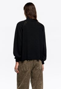 Czarny dziany sweter z wysokim kołnierzem, z raglanowymi rękawami i małymi wycięciami na ramionach. Do kompletu teksturowane oliwkowozielone spodnie.