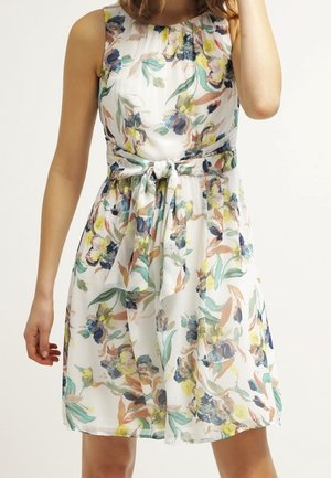 Woman wearing sleeveless white dress with blue, yellow, and green floral pattern and tied waist sash.