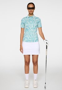 Jasnoniebieska koszulka polo z abstrakcyjnym wzorem, biała spódnica, białe skarpetki i beżowe buty. Trzyma kij golfowy, nosi okulary przeciwsłoneczne.