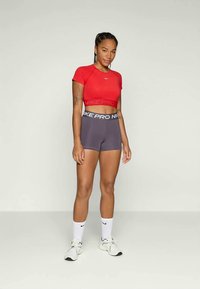 Woman standing wearing red cropped athletic top, gray Nike Pro shorts, white Nike socks, and white sneakers, against plain background.