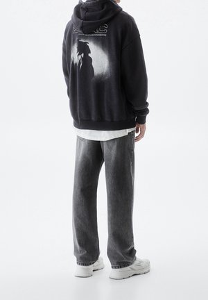 Personne debout, de dos face à la caméra, portant un sweat à capuche noir avec un motif blanc, un jean gris et des baskets blanches sur un fond uni.