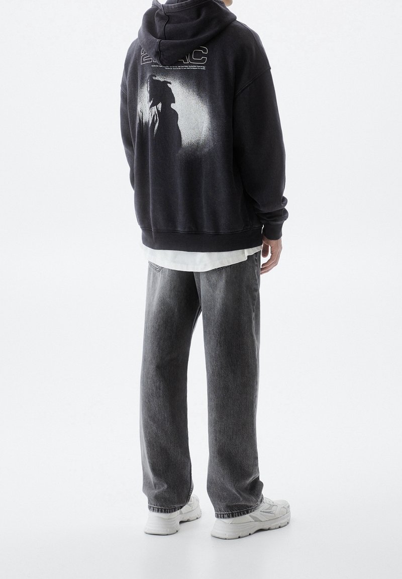 Personne debout, de dos face à la caméra, portant un sweat à capuche noir avec un motif blanc, un jean gris et des baskets blanches sur un fond uni.