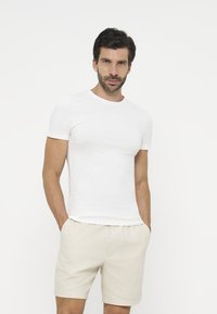 Man with short dark hair and beard wearing a plain white short-sleeve t-shirt and light beige shorts with hands in pockets on white background.