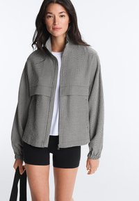 Jeune femme portant une veste grise zippée sur un T-shirt blanc et un short noir, tenant une sangle noire dans sa main gauche.