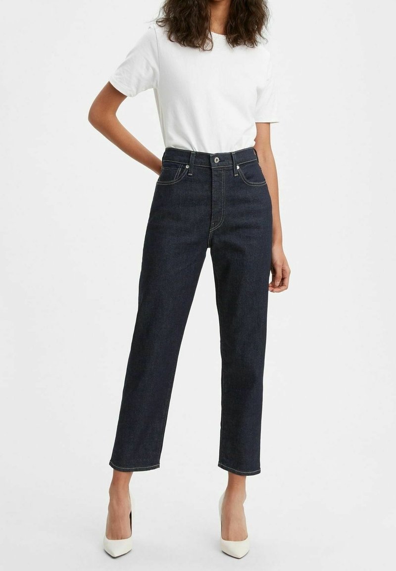 Personne portant un jean bleu foncé taille haute à jambe droite, une chemise blanche à manches courtes et des chaussures blanches à talons hauts, se tenant devant un fond uni.