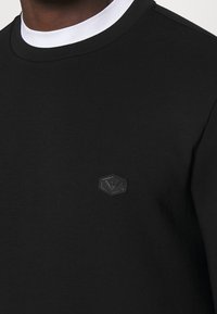 Nära bild av en person som bär en svart sweatshirt med rund halsringning, med en liten hexagonal logotyp på vänster bröst och en vit krage synlig.