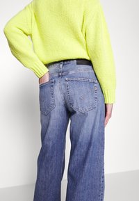 Personne portant un pull en maille jaune vif et un jean bleu taille haute à jambes larges, la main dans la poche arrière, sur un fond uni.