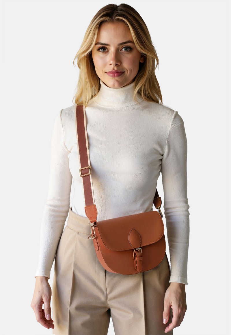Femme portant un pantalon beige et un col roulé blanc, portant un sac bandoulière en cuir marron avec une fermeture à boucle.