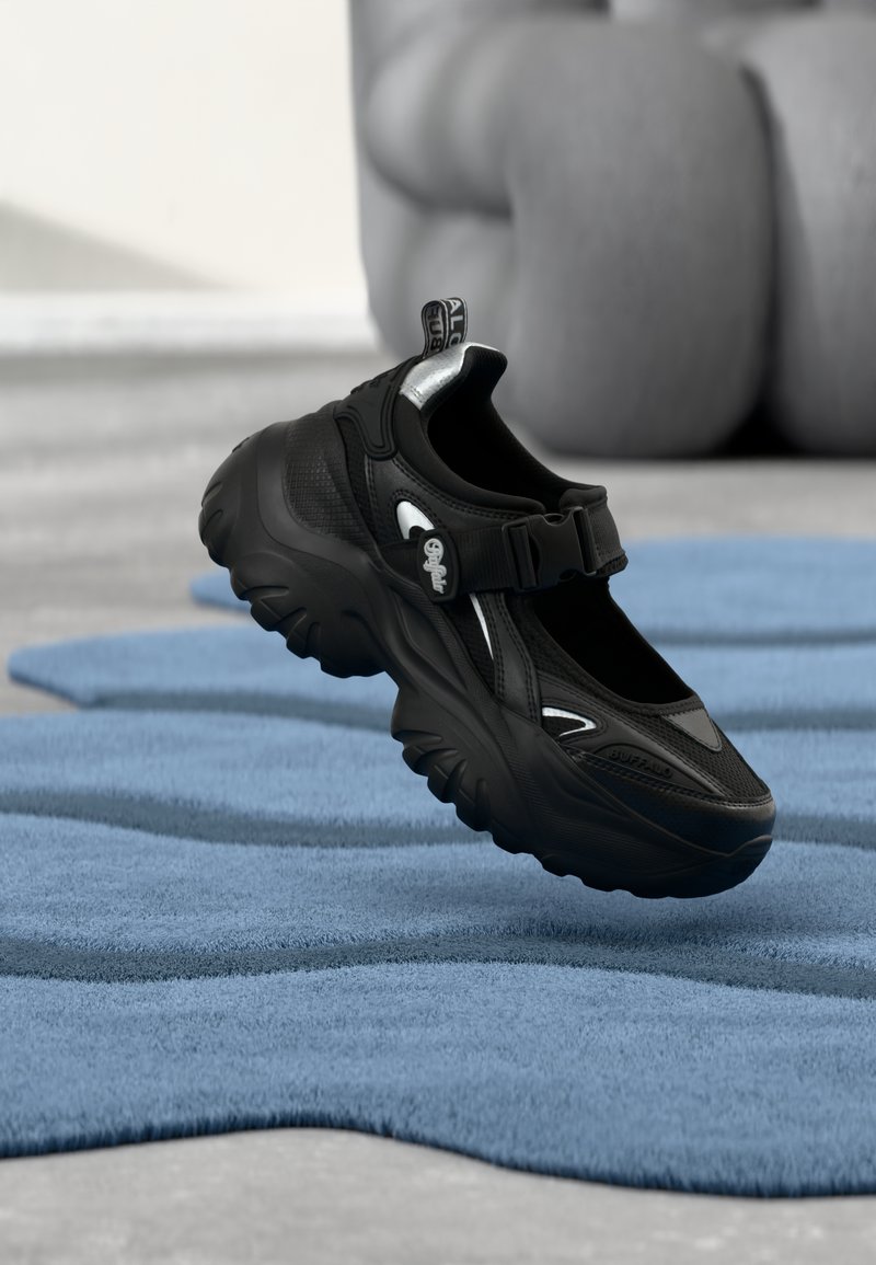 Μαύρο χοντρό αθλητικό παπούτσι με παχιά σόλα και λουράκι Velcro σε μπλε υφασμένο χαλί, με θολό φόντο καθισμένου ατόμου.