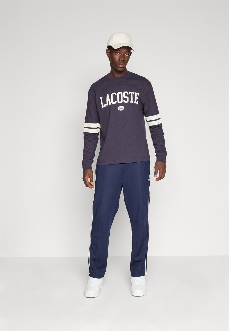 Navy langslaventrui met witte "LACOSTE" tekst en strepen, gecombineerd met marineblauwe trainingsbroek met witte zijkanten en een groen logo. Witte pet en schoenen.
