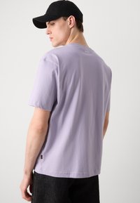 Kortærmet lavendel T-shirt lavet af bomuld, med rund hals, et lille logo på bagsiden og en lige kant. Bæres med mørke bukser.
