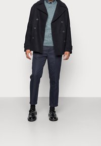 Svart peacoat med breda slag, ljusgrön stickad tröja, marinblå byxor och svarta läderskor med dekorativa detaljer.