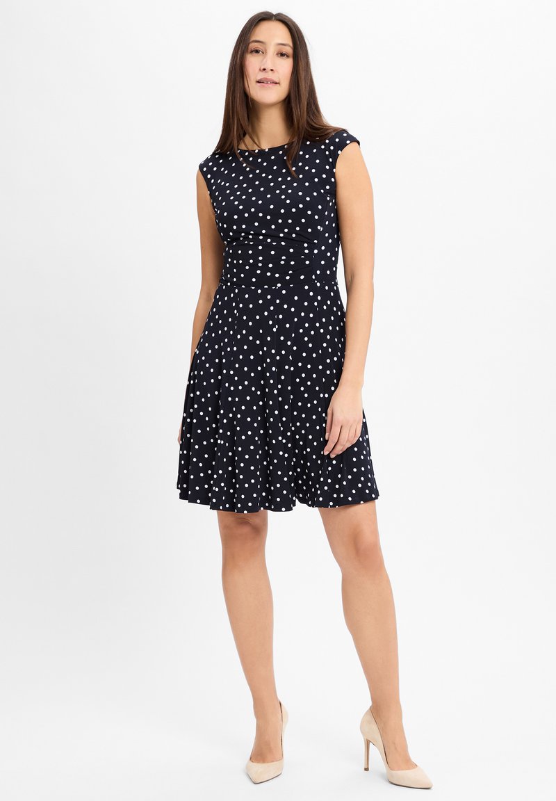 Marineblaues Kleid mit weißen Punkten, kurzen Ärmelchen, tailliertem Oberteil und schwingendem Rock. Glatter Stoff mit leichter Textur.
