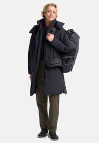 Zwarte geïsoleerde parka met een hoge kraag, gecombineerd met donkere broeken en sneakers. Model draagt een grote donkere rugzak.
