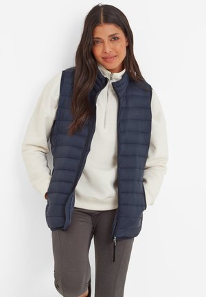 Woman with long dark hair wears a navy quilted vest over a white pullover and gray pants, standing with hands in her vest pockets, smiling slightly.
