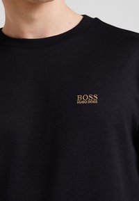 Svart bomullströja med rund hals, prydd med en liten guld broderad "BOSS HUGO BOSS"-logotyp på vänster bröst. Slät textur.