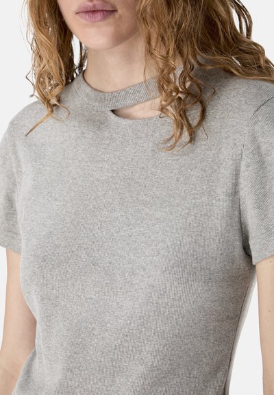 Femme aux cheveux bouclés portant un haut à manches courtes côtelé gris clair avec une petite découpe sous l'encolure.