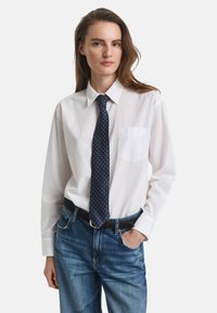 White button-up shirt with a pocket, paired with a dark blue tie featuring a geometric pattern, worn with light blue jeans and a black belt.