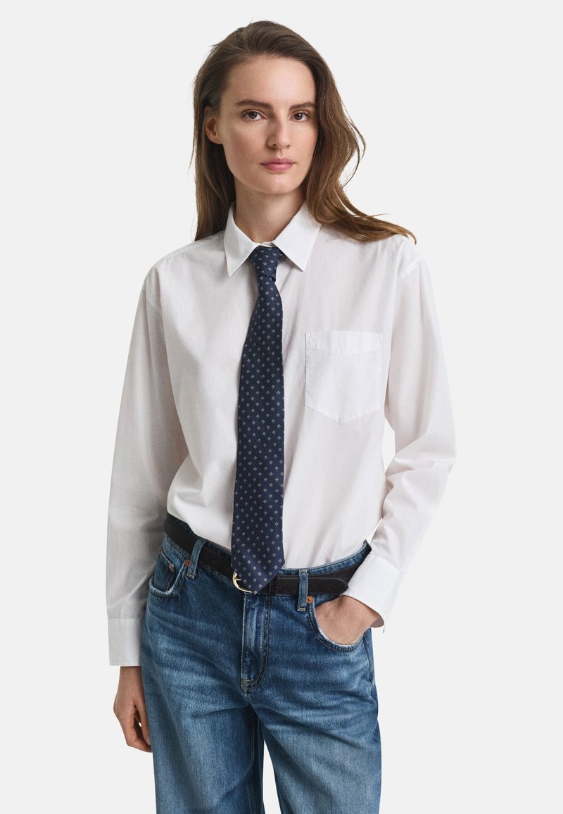 White button-up shirt with a pocket, paired with a dark blue tie featuring a geometric pattern, worn with light blue jeans and a black belt.