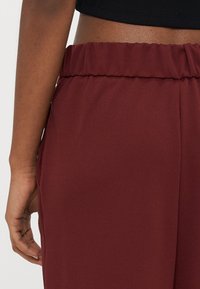 Pantalones color marrón oscuro con una textura suave, que cuentan con una cintura elástica. Se pueden ver bolsillos en las costuras laterales, mostrando un diseño de corte relajado.